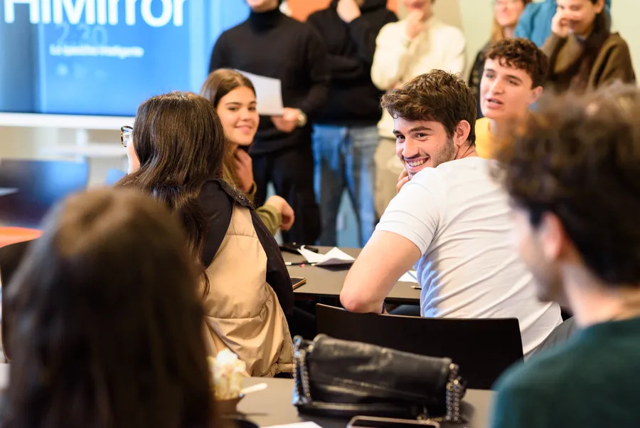 Giovani seduti ai tavoli ascoltano un intervento, davanti a uno schermo